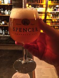 A hand lifts a chalice of beer in toast. The glass bears the name of the Trappist monastery that brews the ale inside: Spencer. The ale is the color of copper and topped with a stiff, high foam.
