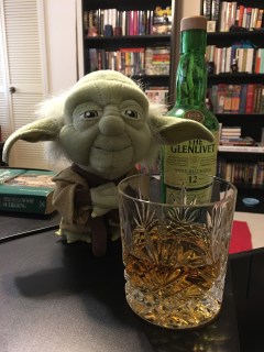 A whisky tumbler with a double measure of The Glenlivet 12-Year-Old sits on a desk before a half-size bottle of same and a stuffed Yoda keeps watch. Background: Bookcases.